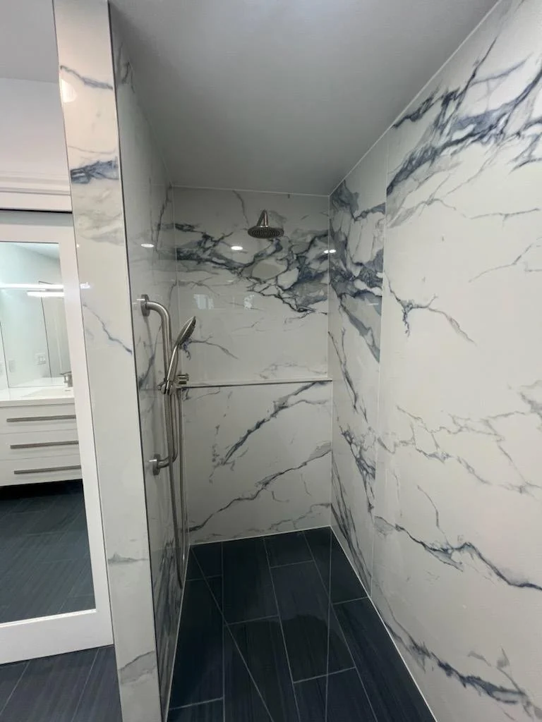 Walk-in shower with marble-pattern tile, window and dark floor.