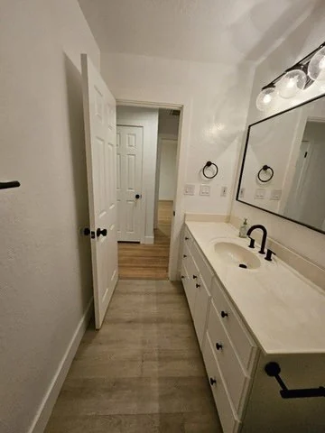 Bathroom with a long white vanity, illuminated mirrors and wood-look floor.