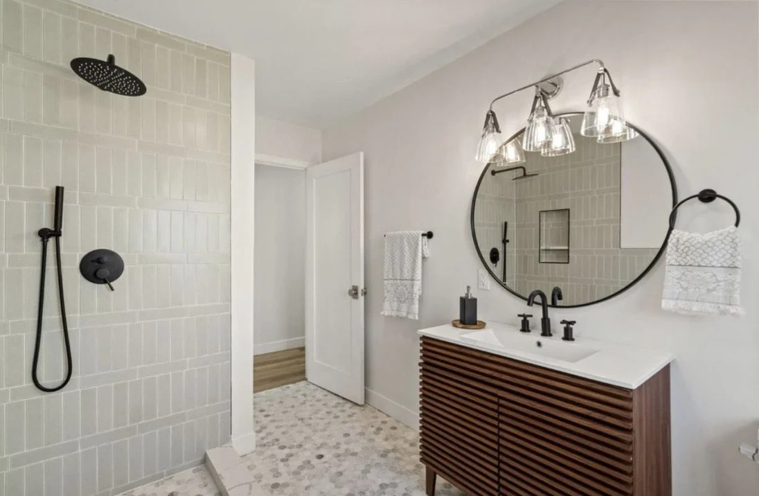 Bathroom with a round mirror, wood vanity, black fixtures and pale shower tile.